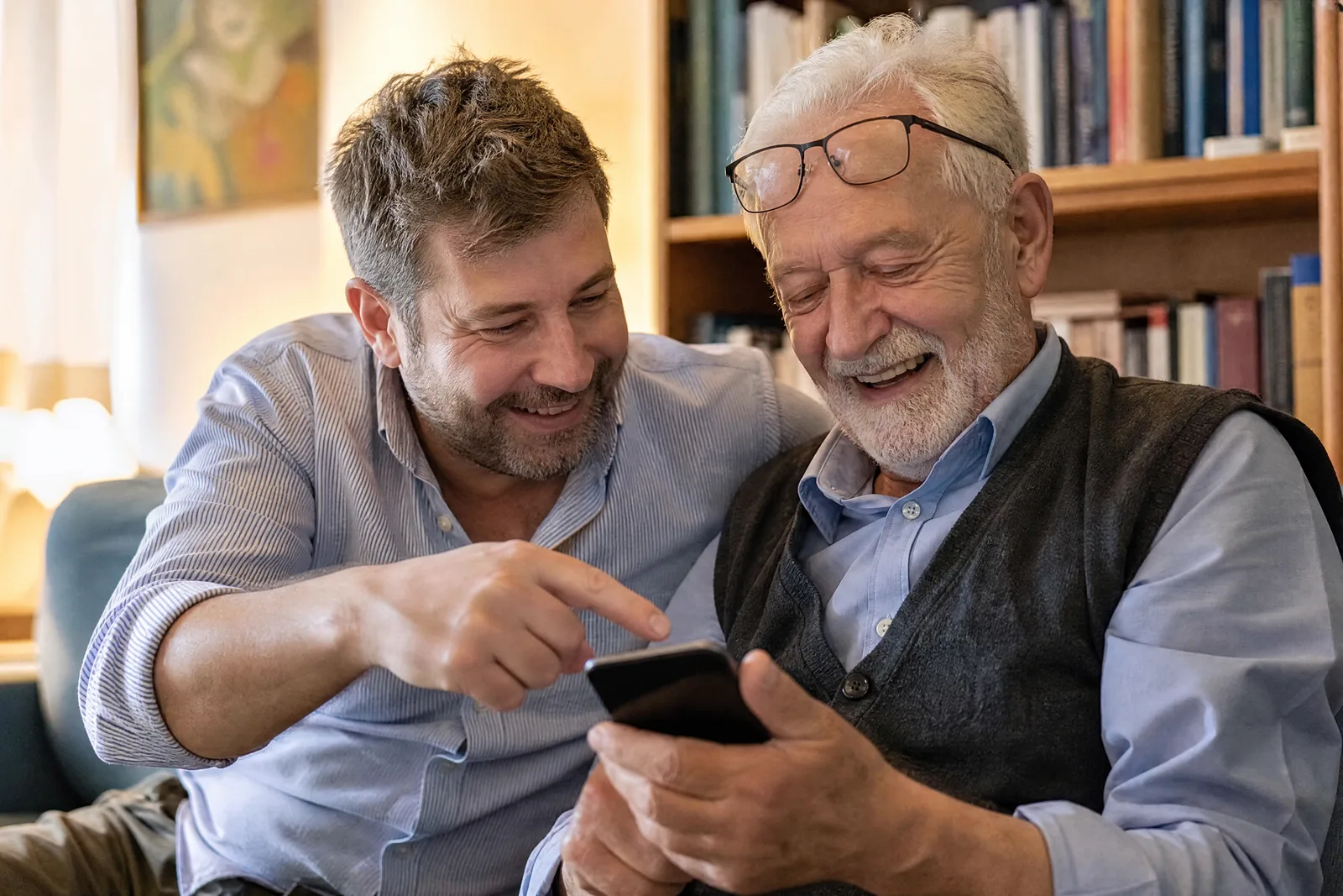 Ein Mann zeigt seinem Vater auf dem Sofa etwas auf dessen Handy