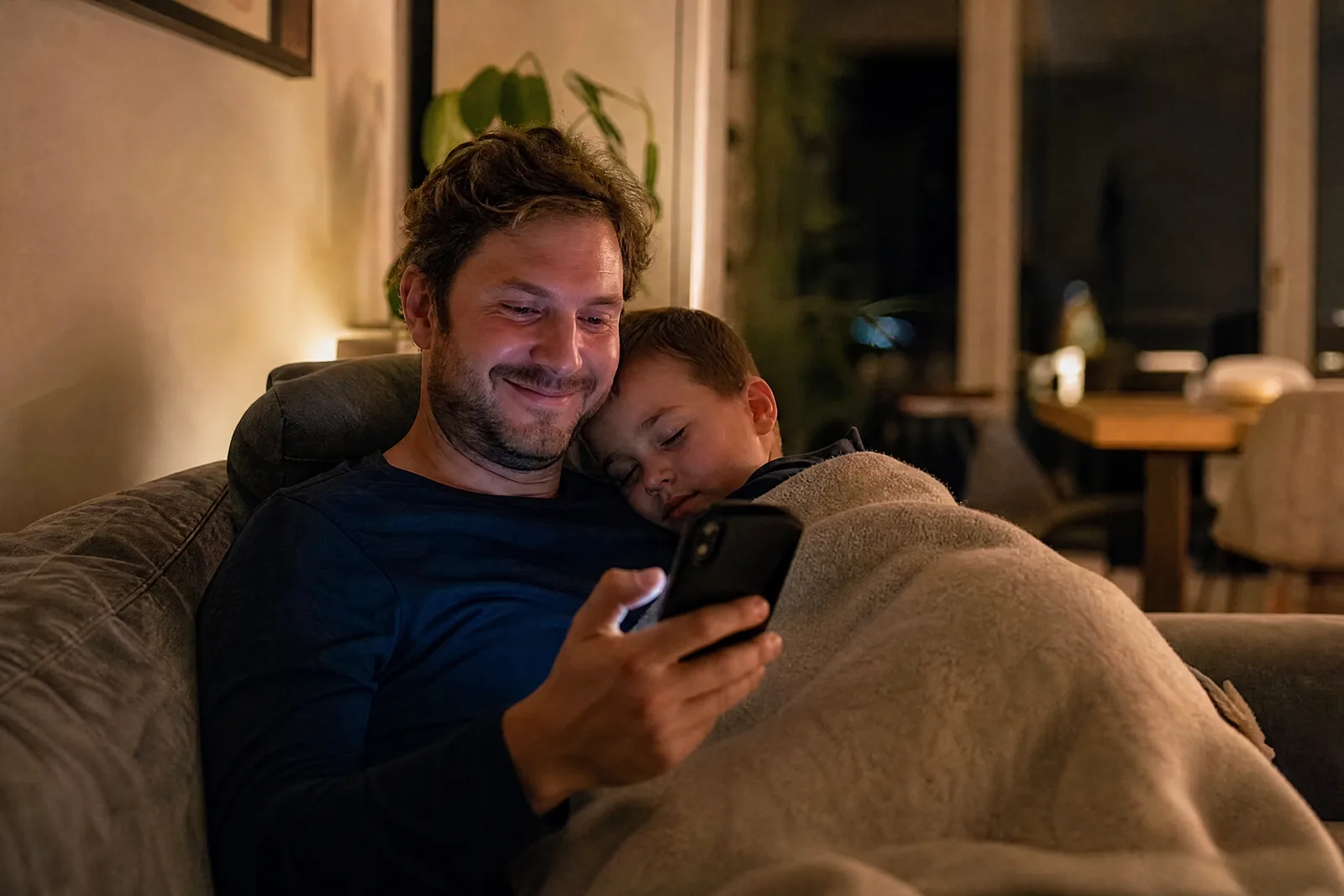 Ein Vater schreibt abends auf dem Sofa eine Nachricht, sein Sohn ist an seiner Schulter eingeschlafen