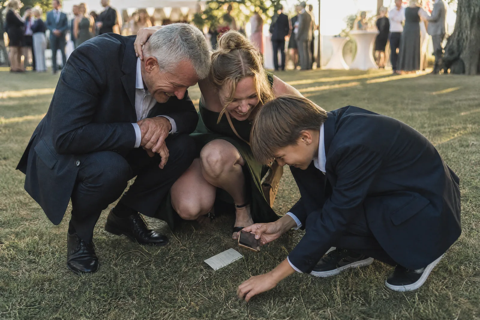Drei Hochzeitsgäste hocken lachend im Gras, um mit dem Handy etwas Kleines zu fotografieren