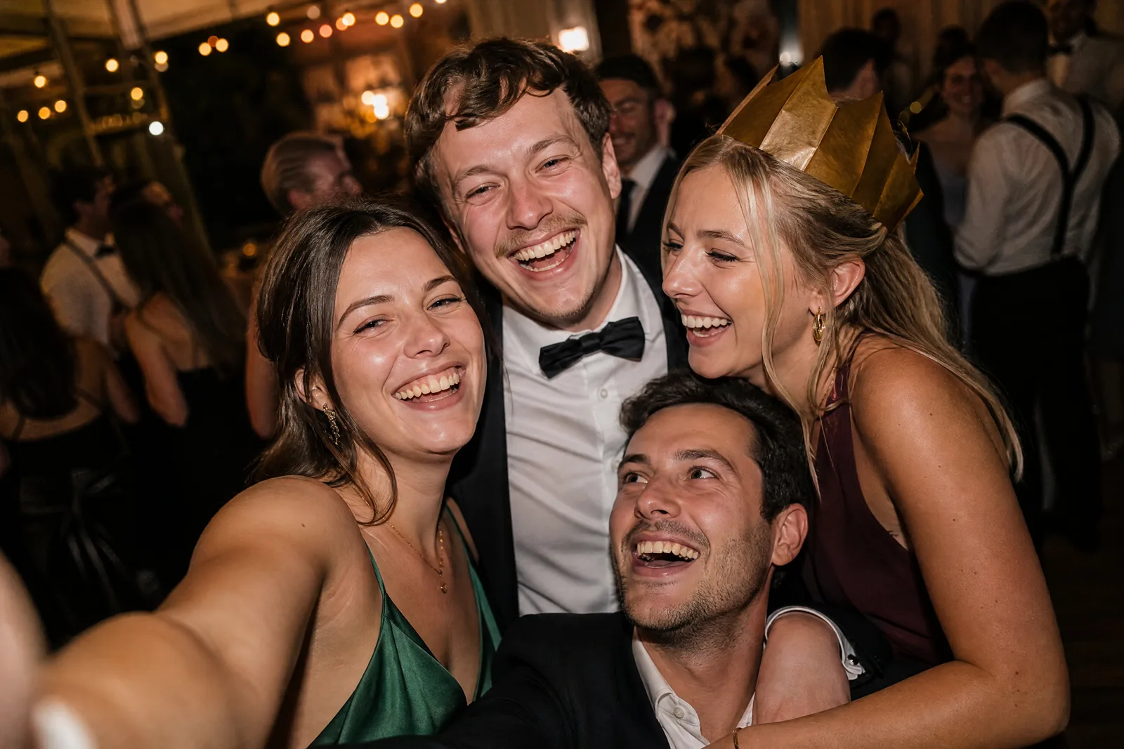 Vier Hochzeitsgäste machen lachend ein gemeinsames Selfie, einer trägt eine schiefe Papierkrone