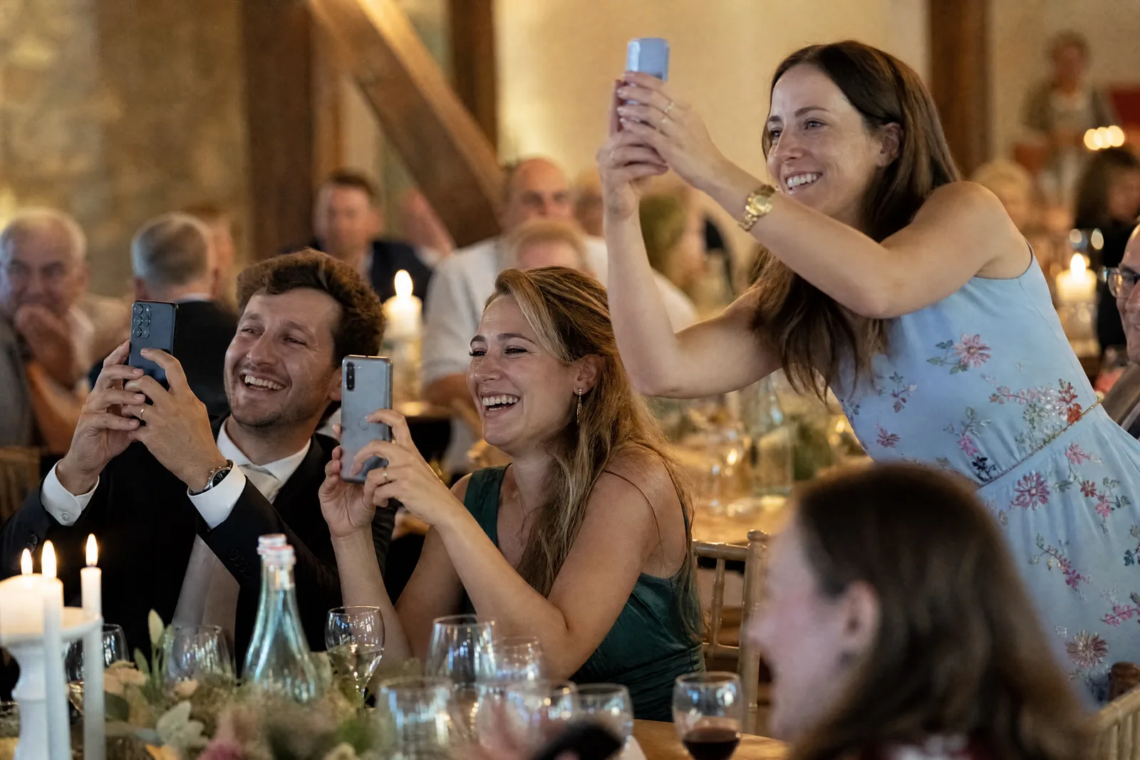 Drei Hochzeitsgäste heben am Tisch ihre Handys, um einen Moment der Feier zu fotografieren