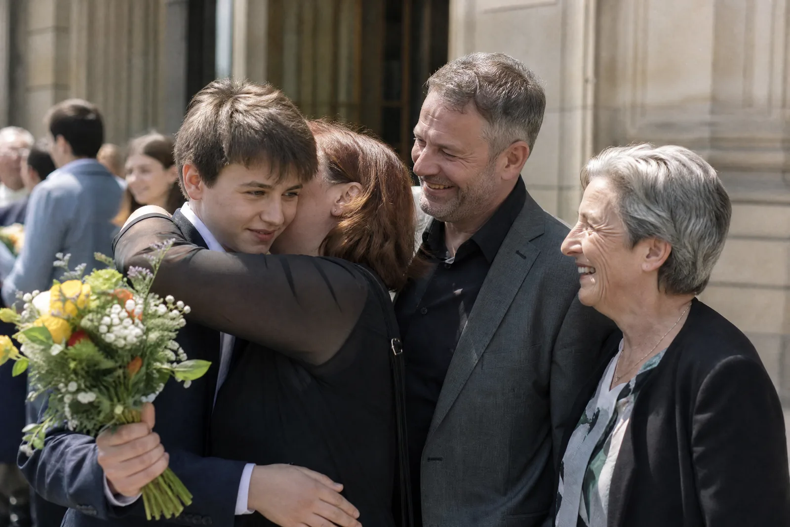 Ein Vierzehnjähriger wird nach der Jugendweihe von seiner Mutter umarmt, mit Blumen in der Hand