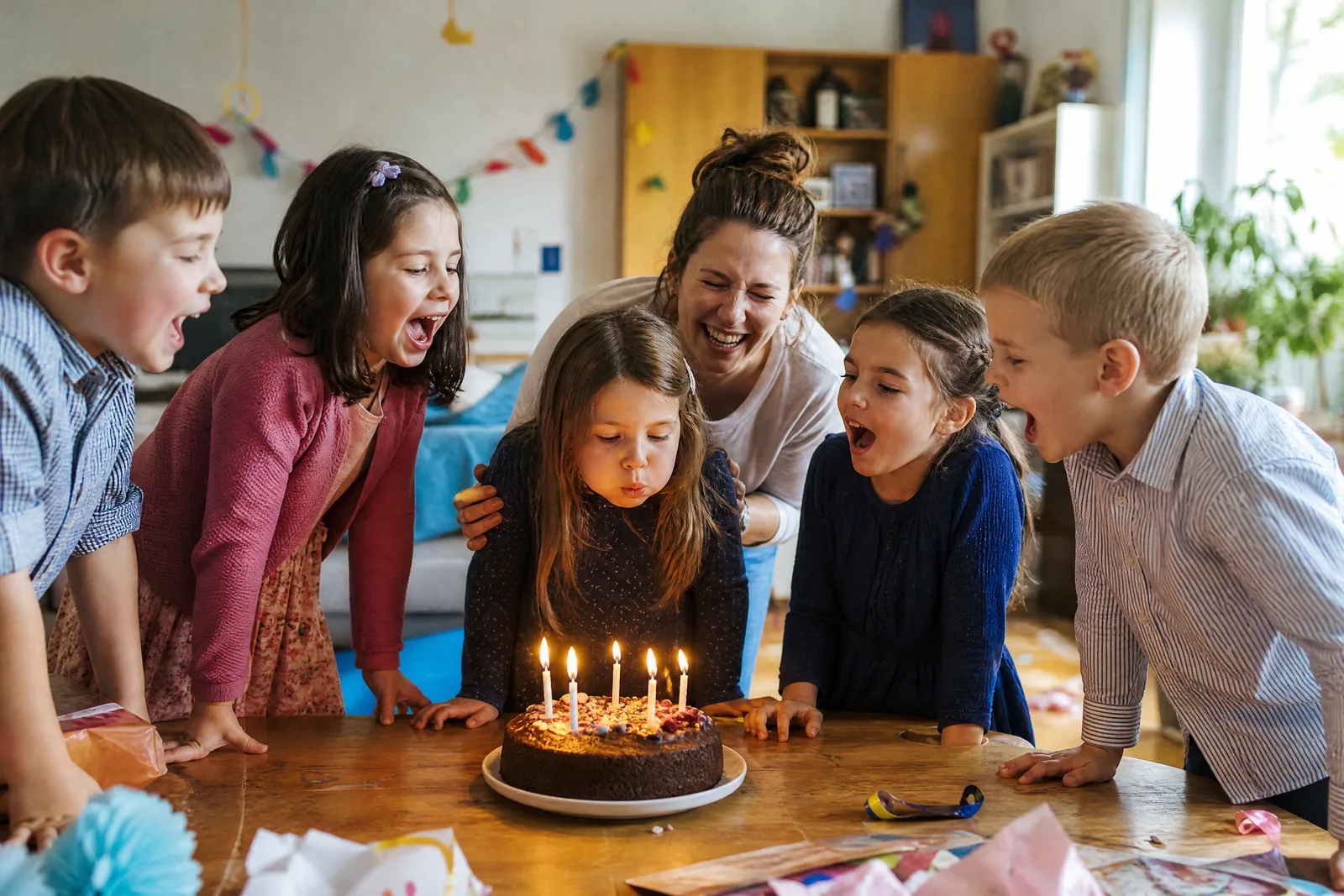 Ein Mädchen pustet die Kerzen auf seiner Geburtstagstorte aus, fünf Kinder und die Mutter jubeln drumherum