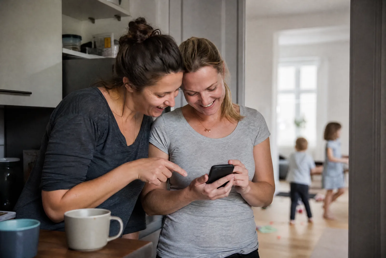 Zwei Mütter schauen gemeinsam lachend auf ein Handy, im Hintergrund spielen Kinder