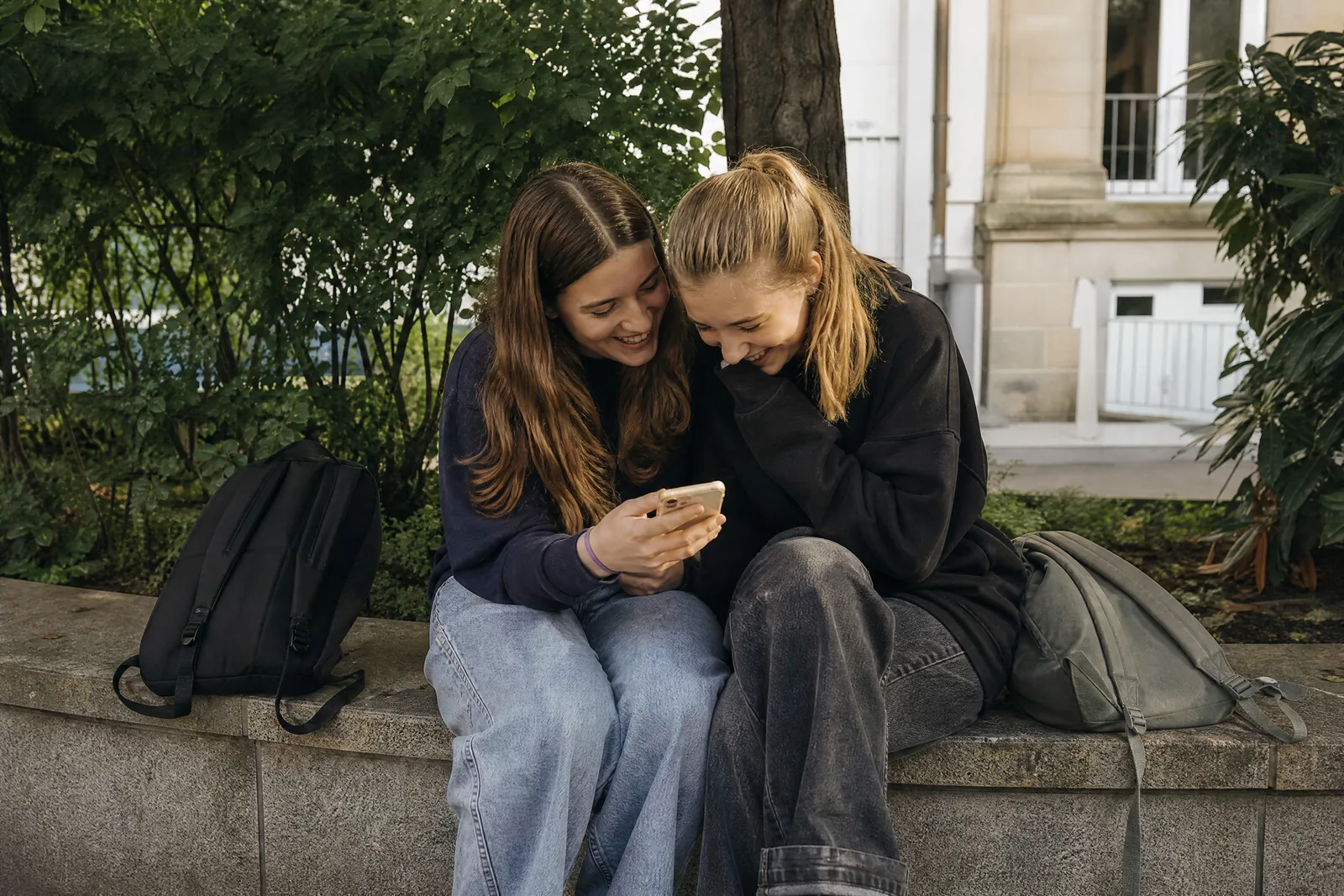Zwei Jugendliche sitzen auf einer Mauer und schauen zusammen lachend auf ein Handy