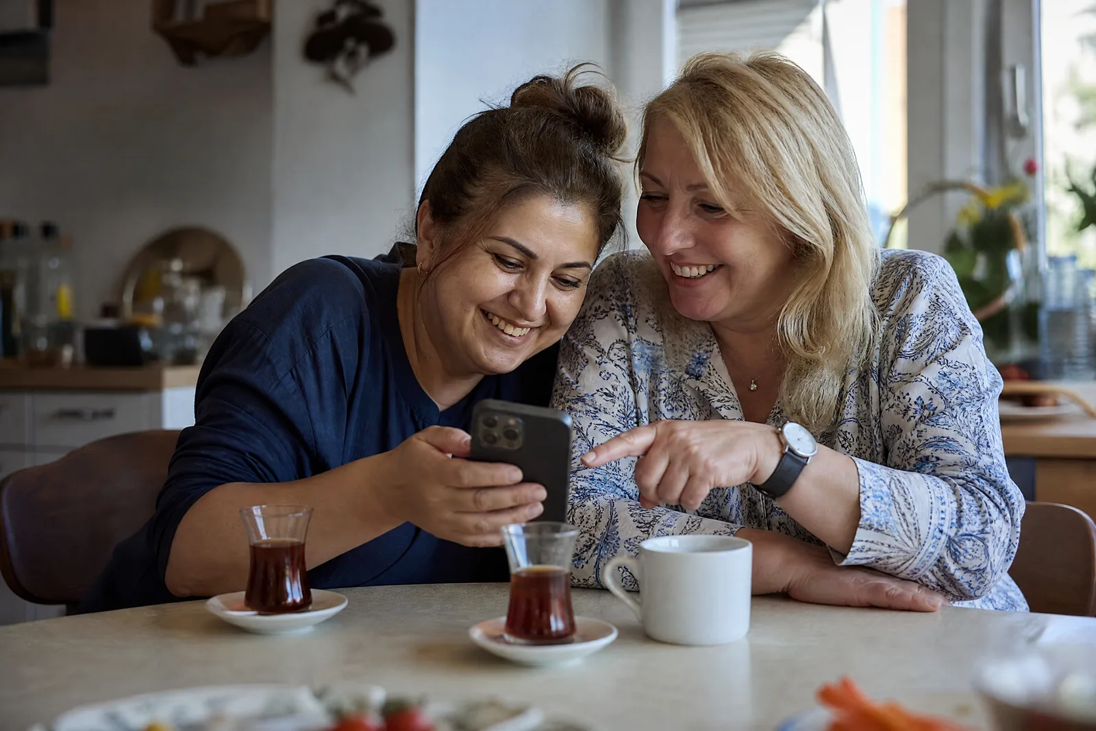 Zwei Mütter sehen sich am Küchentisch gemeinsam eine Einladung am Handy an